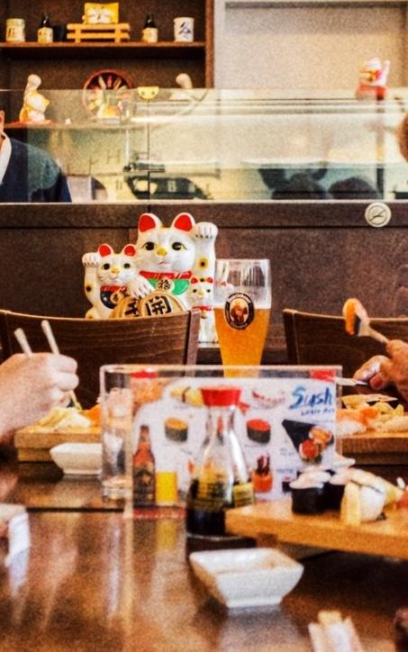 Gäste genießen Sushi an einem Tisch mit Sojasaucenflaschen und einem Glas Bier, während ein Sushi-Koch hinter der Theke arbeitet und dekorative Glückskatzen ausstellt.