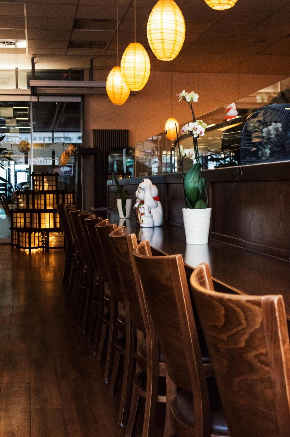 Thekenbestuhlung in einem Sushi-Restaurant mit Holzstühlen, hängenden Laternen, Topfpflanzen und einer ausgestellten Maneki-neko-Statue.