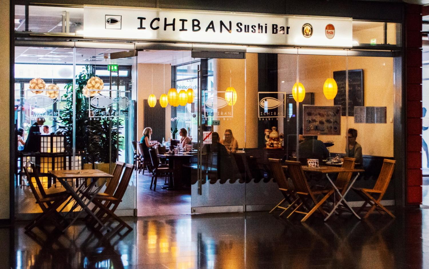 Eingang der Ichiban Sushi Bar in Koblenz mit Glastüren mit dem Logo des Restaurants, Sitzgelegenheiten im Innenraum und warmen Hängelaternen.