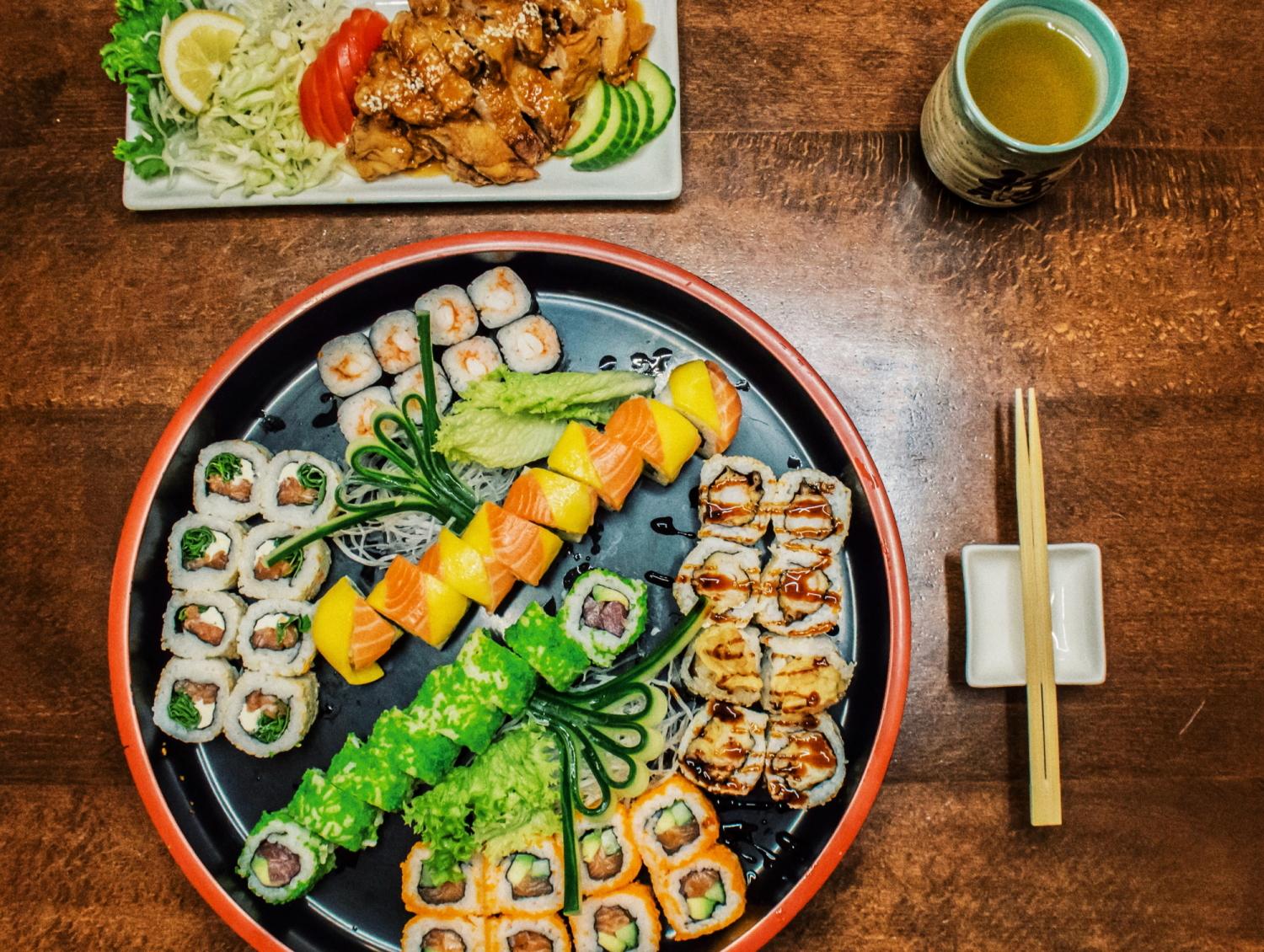 Große Sushi-Platte mit verschiedenen bunten Rollen, serviert mit einer Beilage aus gegrilltem Fleisch und Gemüse und einer Tasse grünem Tee auf einem Holztisch.