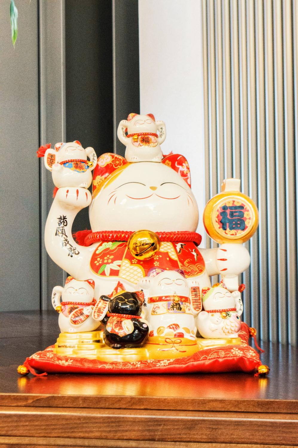 Große Maneki-neko-Statue, umgeben von kleineren Glückskatzen, gekleidet in bunte Dekorationen, ausgestellt auf einem roten Kissen.
